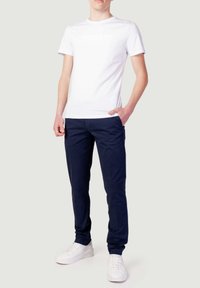 T-shirt bianca in cotone con testo "Morato", abbinata a chinos blu navy e sneakers bianche, che mostrano un outfit casual con un design aderente.