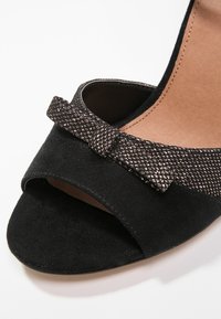 Escarpin ouvert en daim noir avec un nœud métallique texturé en accent sur la bride avant et une doublure intérieure lisse.