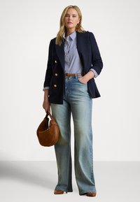 Sieviete valkā tumši zilu žaketi, svītrainu kreklu, plataus kāju džinsus, brūnu jostu un kurpes, turot rokā brūnu zamšādas roku somu, viena roka ielikta kabatā.