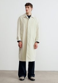 Νέος άνδρας που στέκεται φορώντας ένα μακρύ μπεζ παλτό trench, μαύρο παντελόνι και γκρι παπούτσια, μπροστά σε απλό λευκό φόντο.