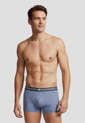 Paitaton mies, jolla on lyhyet ruskeat hiukset, siniset bokserit valkoisilla pilkuilla ja mustalla vyötärönauhalla, katsoo hieman oikealle.