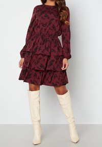 Burgundy mekko, jossa on musta kukkakuosi, pitkät hihat, kerroksellinen muotoilu ja joustava vyötärö. Yhdistetty polveen ulottuviin luonnonvalkoisiin saappaisiin.