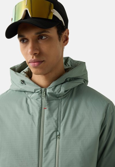 Young man wearing a light green hooded jacket, black cap, and reflective yellow sports sunglasses resting on the cap brim.