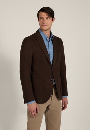 Hombre con una chaqueta marrón oscura, camisa azul de botones y pantalones beige, de pie frente a un fondo claro y liso.