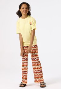 T-shirt en coton jaune à manches courtes, associé à un pantalon évasé à motifs zigzag rouge et orange. Le mannequin porte des sandales noires à enfiler.