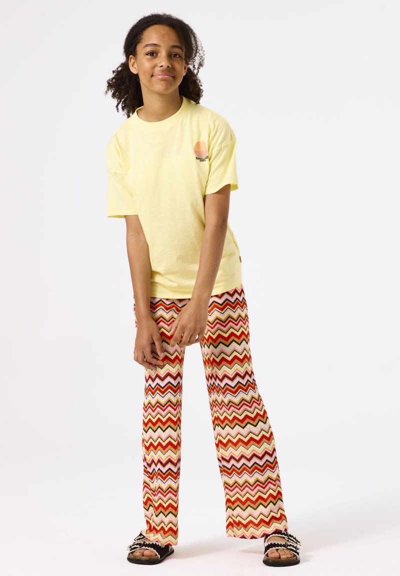 T-shirt en coton jaune à manches courtes, associé à un pantalon évasé à motifs zigzag rouge et orange. Le mannequin porte des sandales noires à enfiler.