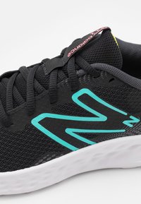 Zapatilla deportiva negra con parte superior de malla, acentos turquesas, patrón circular texturizado y suela blanca. Presenta cordones negros y una etiqueta de marca.