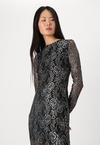 Robe en dentelle noire avec de longues manches transparentes et des motifs floraux en fil argenté. Présente une silhouette ajustée et un col rond.