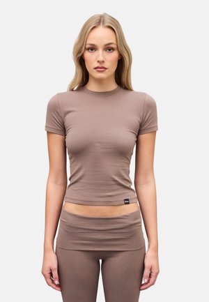 Mujer con cabello largo y rubio, vestida con una camiseta ajustada de manga corta color topo y leggings a juego de cintura alta, sobre un fondo liso.