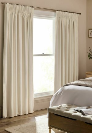 Helles Schlafzimmer mit geschlossenen cremefarbenen Vorhängen über einem Fenster, einem Bett mit weißer Bettwäsche und einer gepolsterten Bank mit einem offenen Buch.