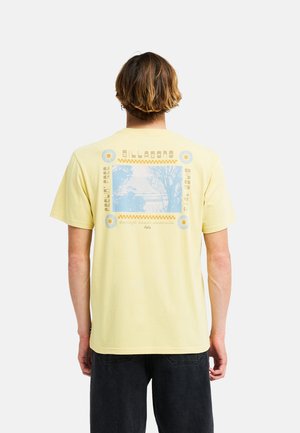 Uomo che indossa una t-shirt giallo chiaro con un design grafico blu e giallo e testo sulla schiena, abbinata a pantaloni neri.