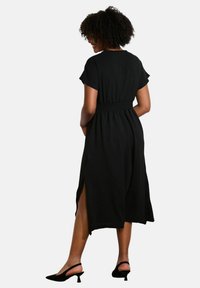 Black, knee-length dress with short sleeves, elastic waistband, and side slits. Fabric appears lightweight and smooth. Worn with black heels.