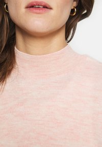 Naisen alavartalo, kasvojen alaosa ja kaula, joissa on kultaiset rengaskorvakorut ja vaalea vaaleanpunainen ribbikuvioinen poolopaita.