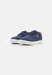 Navy canvas sneakers with white rubber soles, featuring brown leather accents, six eyelets, and flat laces. Textured surface and minimalist design.