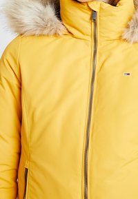 Geel gewatteerde jas met een nepbont capuchon, ritssluiting aan de voorkant en zijzakken. Heeft een klein logo op de linkerborst.