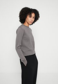 Maglione grigio chiaro lavorato a maglia con collo rotondo, polsini e orlo a costine. Indossato con pantaloni neri, che mettono in risalto un design aderente.