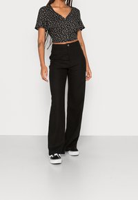 Zwarte cropped top met wit bloemenpatroon, gecombineerd met hoge tailles zwarte wijde broek en zwarte sneakers. Soepele stofstructuur.