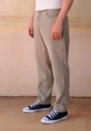 Person trägt beige Cordhosen und schwarze Segeltuch-Sneaker mit weißen Schnürsenkeln, steht auf einem strukturierten Boden vor einer zweifarbigen Wand.