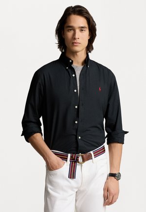 Black button-down shirt with a collar, worn over a gray t-shirt. Paired with white pants and a multicolored striped belt.