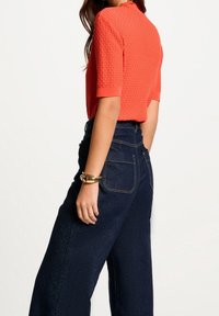Koraltowy top z teksturowanego dzianiny z krótkimi rękawami, zestawiony z ciemnoniebieskimi jeansami o szerokich nogawkach i wysokim stanie. Na nadgarstku złota bransoletka.