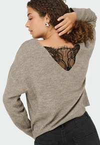 Lichtbeige trui met een losse pasvorm, V-hals en kanten paneel aan de achterkant. Gestructureerde stof, lange mouwen, gecombineerd met zwarte broek.