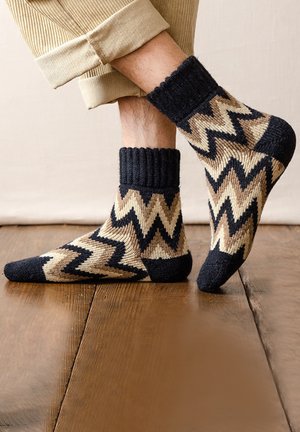 Knit socks featuring a zigzag pattern in navy, cream, and beige. Cuffed design with a textured finish. Positioned on a wooden floor.
