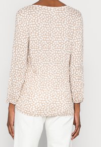 Blusa beige con motivo a mosaico, maniche a tre quarti, scollo rotondo e tessuto texture. La vista posteriore mostra una vestibilità ampia.