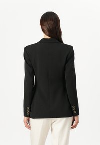 Blazer nero con spalle strutturate, dotato di una singola fessura posteriore e polsini con bottoni dorati, materiale testurizzato e vestibilità sartoriale.