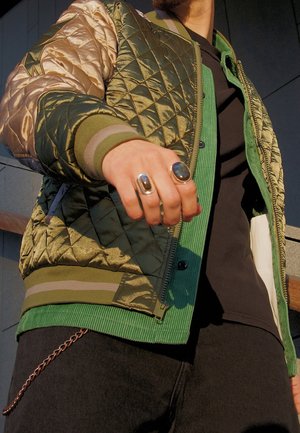 Mężczyzna w oliwkowo zielonej pikowanej kurtce i zielonej koszuli, wyciągający rękę do przodu z dwoma dużymi pierścieniami, łańcuch zwisający z czarnych spodni.