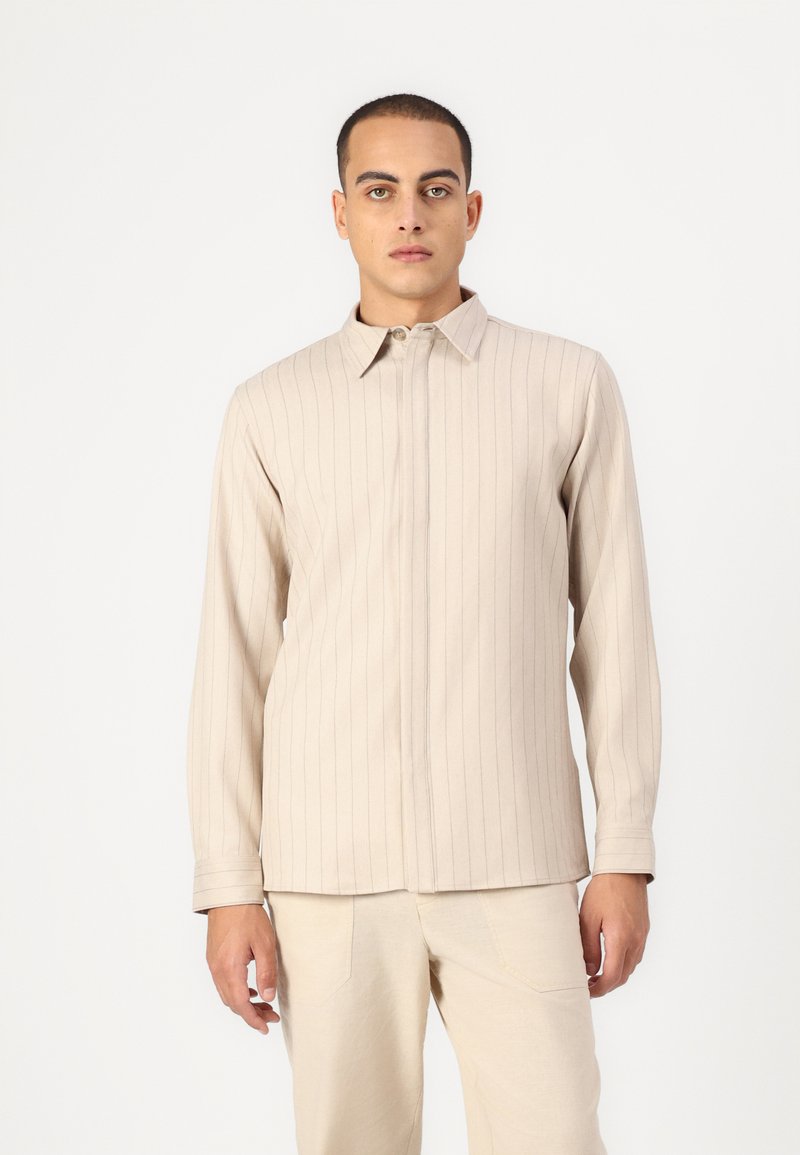 Beige košeľa s dlhým rukávom, vertikálnymi pruhmi, gombíkovým golierom a centrálnym zipsom. Materiál vyzerá hladko a má štruktúrovaný strih.