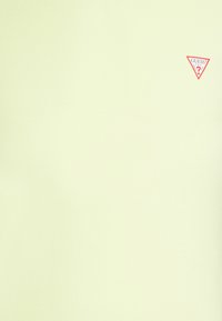 Světle žluté tričko vyrobené z jemné látky, s malým červeným logem Guess na levém horním rohu. Jednoduchý design, žádné další vzory nebo detaily.