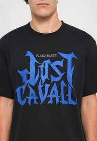 Fekete pamut póló, nagy kék grafikai szöveggel, textúrázott részletezéssel, amely a "FIRED EARTH" feliratot tartalmazza a "JUST CAVALLI" felett. Állítható kerek nyakkivágás.