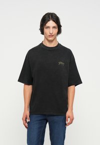 Svart oversized t-skjorte i bomull med rund hals og korte ermer, med et lite grønt grafisk design på venstre brystområde.