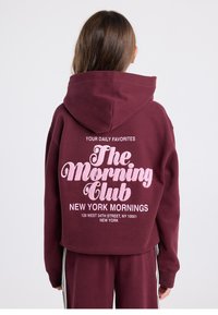 Viininpunainen huppari, jossa on vaaleanpunainen kimaltava grafiikka takana, jossa lukee "The Morning Club" ja "New York Mornings". Huppari on rento ja siinä on huppu.