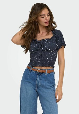 Jeune femme aux cheveux bruns lâchés, portant un top court à pois bleu marine et un jean bleu taille haute avec une ceinture marron.