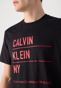 Czarny bawełniany T-shirt z wyraźnym czerwonym napisem: "CALVIN KLEIN" oraz "NY", z dolną linią informującą "Calvin Klein Established 1968 NYC."