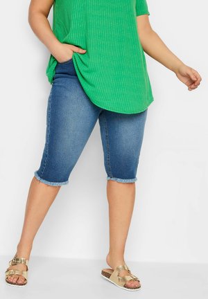 Kvinde iført midt-lange denimshorts med frynset kant, en grøn ribbet skjorte og gyldne sandaler med spænde, stående mod en ensfarvet hvid baggrund.