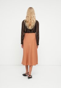 Bruine satijnen blouse met lange mouwen, gecombineerd met een midi-lengte, vloeiende, lichtbruine rok. Zwarte haksandalen maken de outfit compleet.