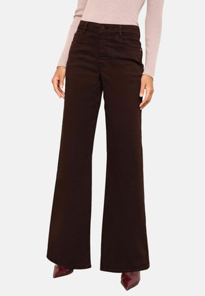 Braune weit geschnittene Jeanshosen mit genähten Akzenten, vorderen Taschen und einem Knopfverschluss. Getragen mit einem hellen Oberteil und spitz zulaufenden Schuhen.