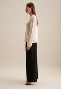 Beige gebreide trui met een v-hals en geribbelde manchetten, gecombineerd met zwarte broek met wijde pijpen. Model staat zijwaarts tegen een neutrale achtergrond.