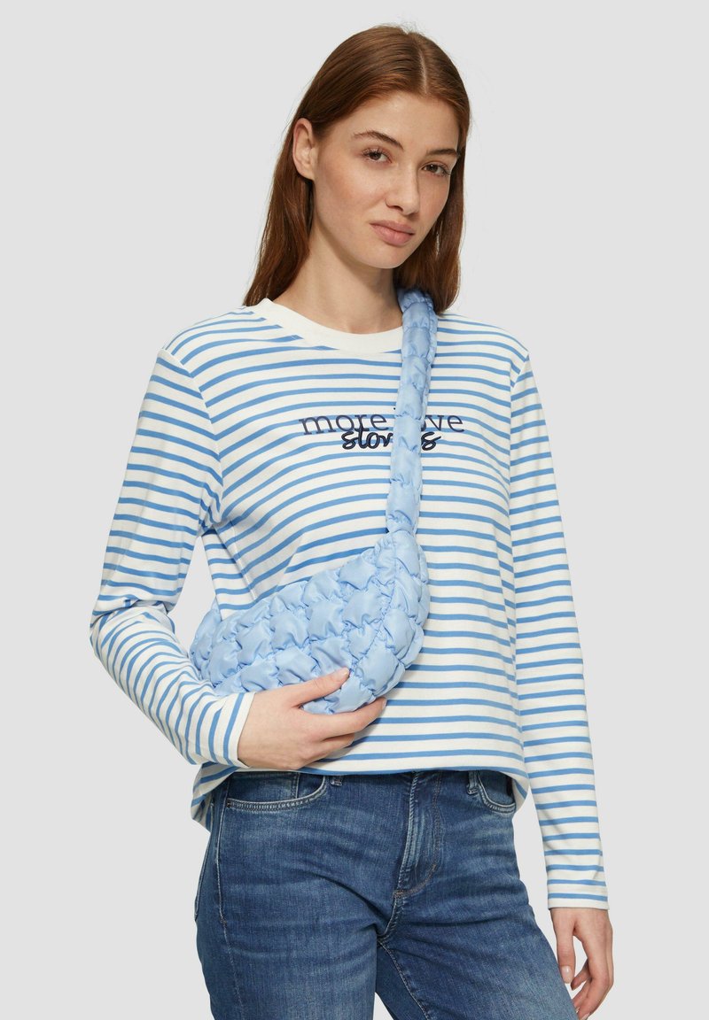 Helleblaue, gepolsterte, gewebte Umhängetasche mit einer strukturierten Oberfläche. Das Modell trägt ein gestreiftes Langarmshirt mit dem Aufdruck "mehr Liebesgeschichten".