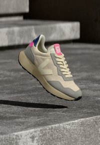 Μπεζ αθλητικό παπούτσι με υφασμάτινο επάνω μέρος, γκρι λεπτομέρειες, ροζ και ναυτικό μπλε ταμπελάκια στη φτέρνα και ανοιχτόχρωμη σόλα. Διαθέτει εμφανές λογότυπο στη γλώσσα.