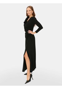 Czarna sukienka z długim rękawem z marszczonym gorsetem i kwiatowym akcentem, z wysokim rozcięciem po lewej stronie, noszona z czarnymi szpiczastymi szpilkami.