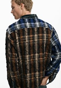 Plaid shirt with a mixed color palette of blue, brown, and cream. Features a textured, patchwork design with frayed edges and a collar.