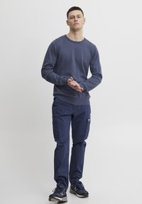 Pulover lung de culoare albastru marin cu manșete cu tiv, asortat cu pantaloni cargo albastre, cu buzunare laterale. Pantofi sport negri completează ținuta.