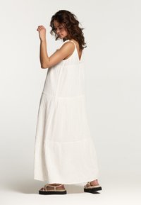 Robe maxi en tissu léger et blanc avec une finition texturée. Caractérisée par un dos en V, des fines bretelles et un design à volants. Sandales légères présentées.