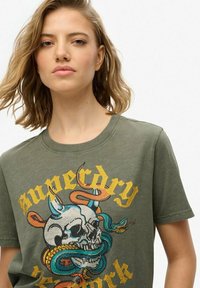 Camiseta verde oliva con cuello redondo, presenta un gráfico detallado de un cráneo entrelazado con serpientes coloridas y el texto "Superdry" en amarillo.