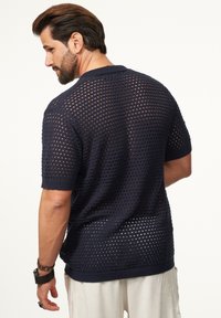 Marineblaue, kurzärmlige Strick-Polohemd mit einem perforierten Mesh-Design, ausgestattet mit gerippten Bündchen und einem Kragen. Ansicht von hinten.