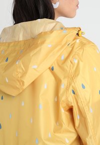 Gul regnjacka med huva, med ett mönster av vattendroppar i ljusblått och vitt. Tillverkad av glänsande, lätt material.