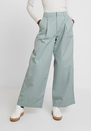 Onderlichaam van een persoon die lichtgroene wijde broek draagt, een wit geribd topje en witte sneakers met bruine zolen, handen in de zakken.